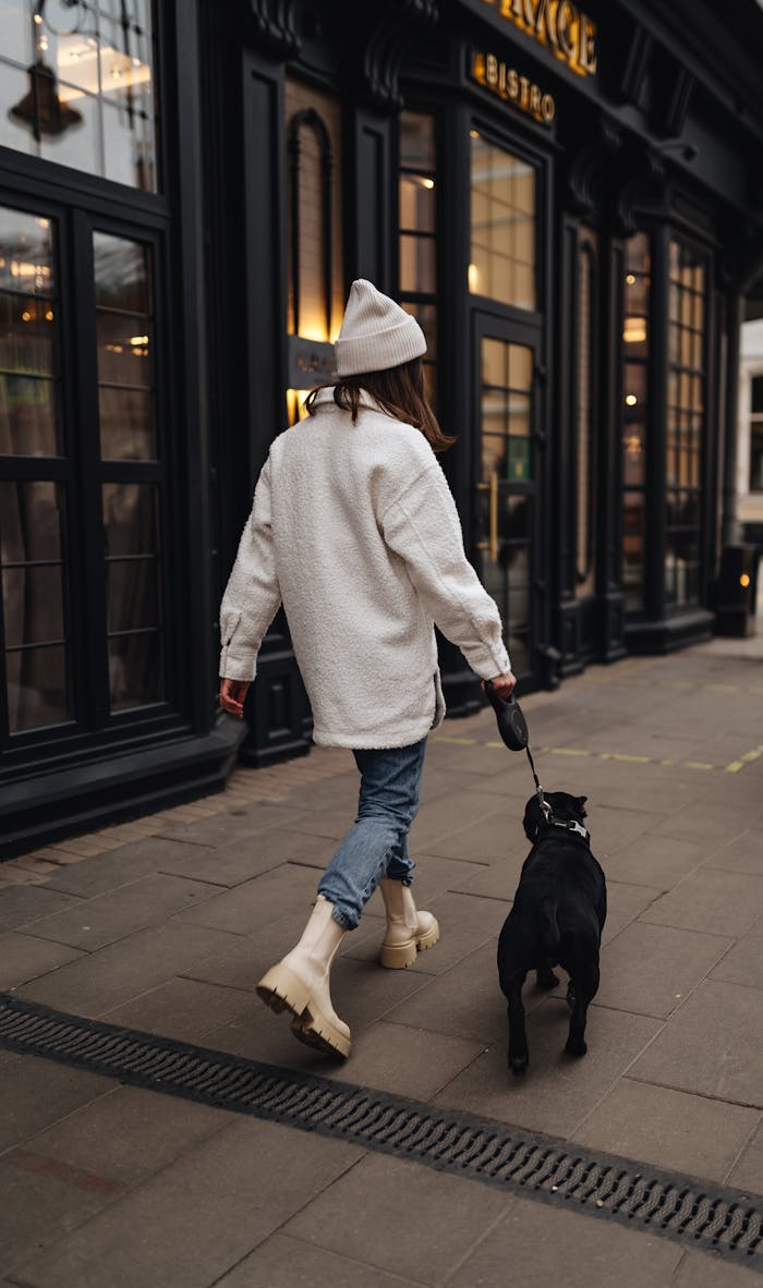 A woman in a white coat and hat walks her dog on a city sidewalk past a bistro.